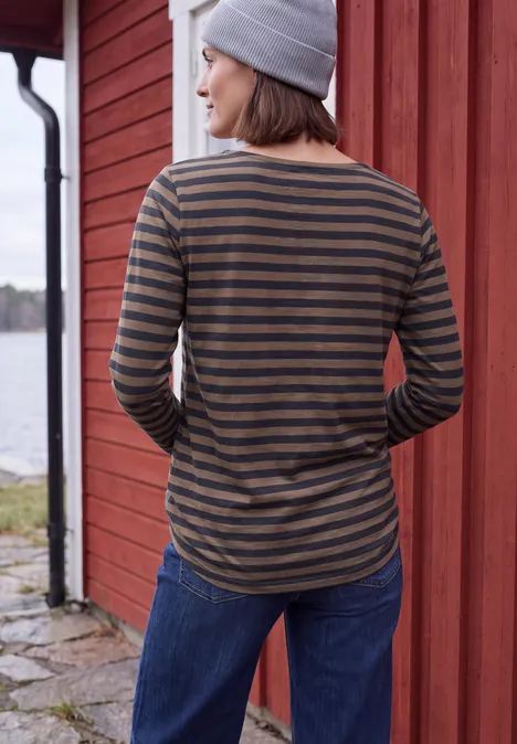 Femme de dos près d’une cabane rouge, pull rayé, bonnet gris et jean, au bord de l’eau.