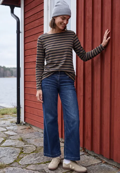 Femme en pull rayé, jean et bonnet, souriante, appuyée contre un mur de bois rouge au bord d'un lac.