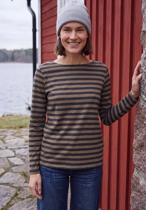 Femme en tee-shirt rayé, jean et bonnet gris appuyée sur un mur rouge près de l’eau.