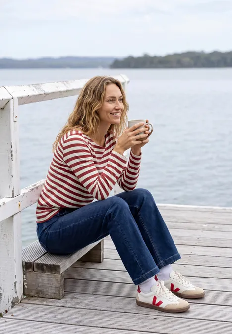 Une femme souriante sur une jetée boit dans une tasse devant l'eau calme.