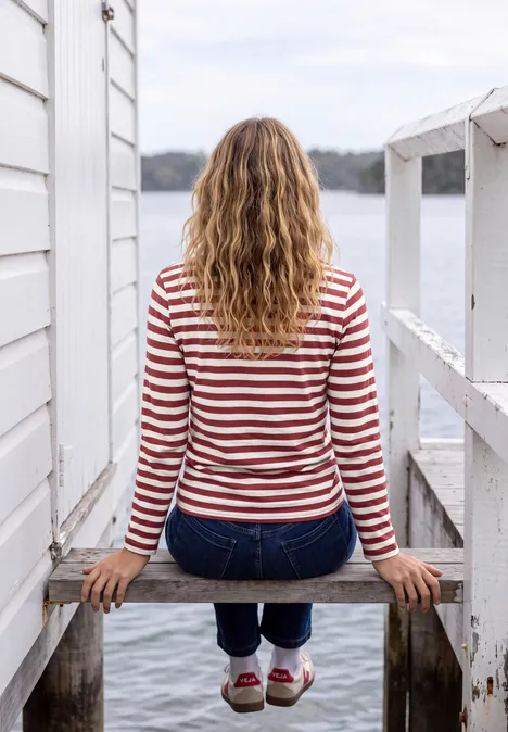 Une femme en marinière rouge et blanche est assise sur un ponton en bois, face à l'eau.