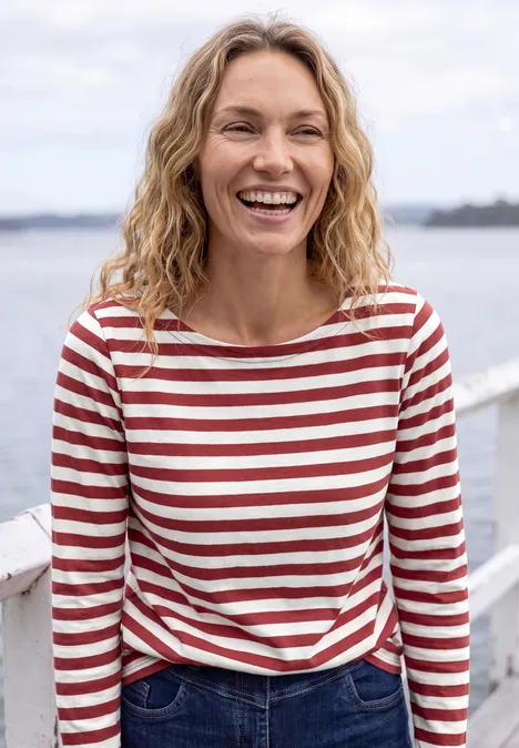 Femme souriante en marinière rouge et blanche, en bord de mer et ciel clair.