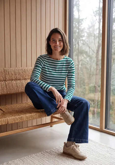 Een lachende vrouw in een gestreepte trui en jeans zit op een houten bank in een lichte kamer.