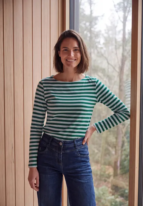 Een glimlachende vrouw in groen-zwarte gestreepte trui en spijkerbroek staat voor een houten wand met boszicht.