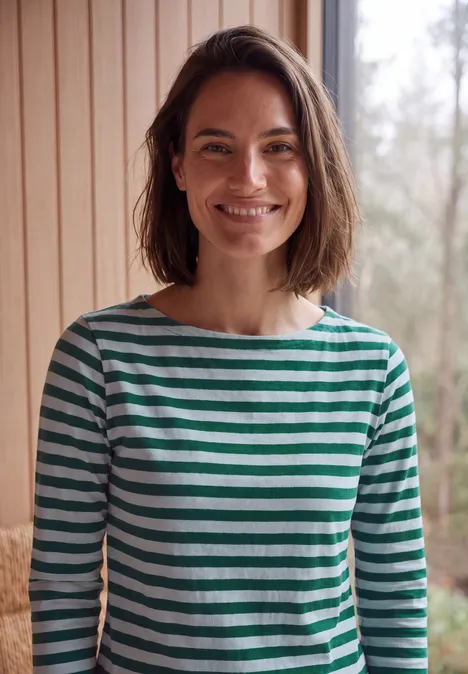 Een vrouw in een groen-wit gestreept shirt lacht voor een licht houten interieur.