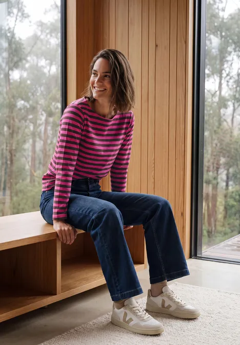 Een vrouw zit glimlachend op een houten bank, draagt een gestreepte trui, jeans en sneakers bij grote ramen.