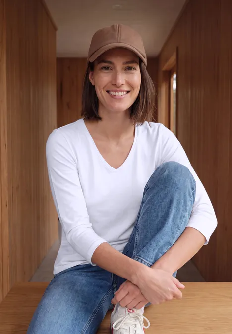 Une femme souriante en jean, haut blanc et casquette, assise devant des parois en bois.