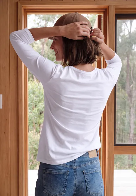Femme de dos en t-shirt blanc et jean, ajuste ses cheveux courts dans une pièce lumineuse avec grandes fenêtres.
