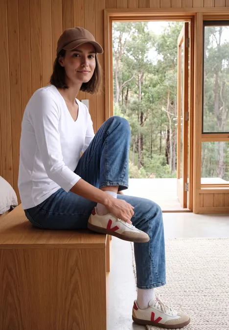 Jeune femme dans une pièce en bois lumineux, noue ses baskets, porte jean et casquette.