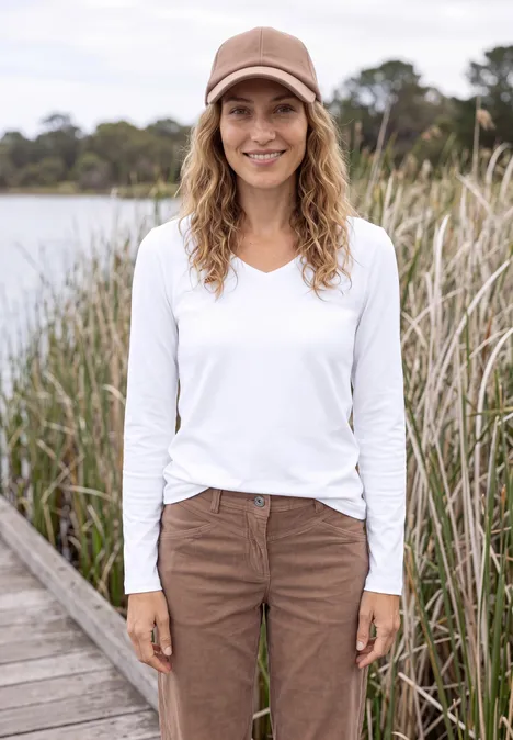 Femme souriante en casquette marron, t-shirt blanc à manches longues et pantalon marron sur un ponton près d’un étang.