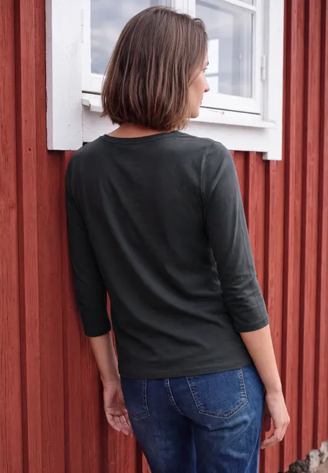 Une femme vue de dos porte un haut noir à manches trois-quarts et un jean, adossée à un mur en bois rouge.