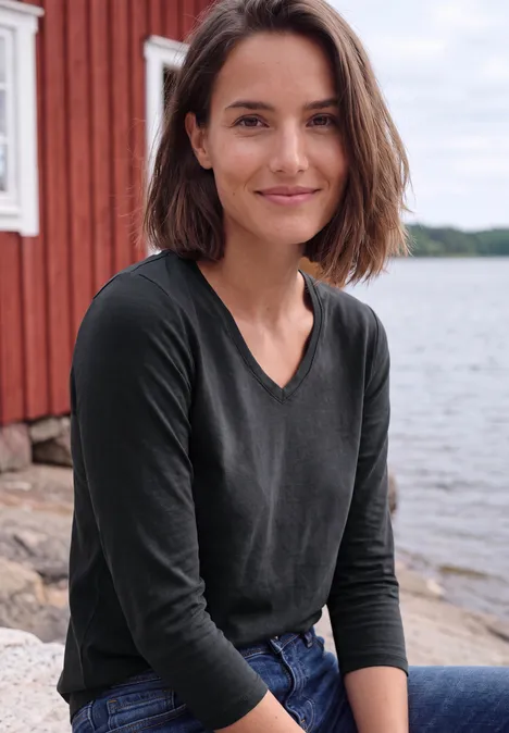 Une femme souriante est assise au bord de l'eau, devant une maison en bois rouge sur un lac calme.