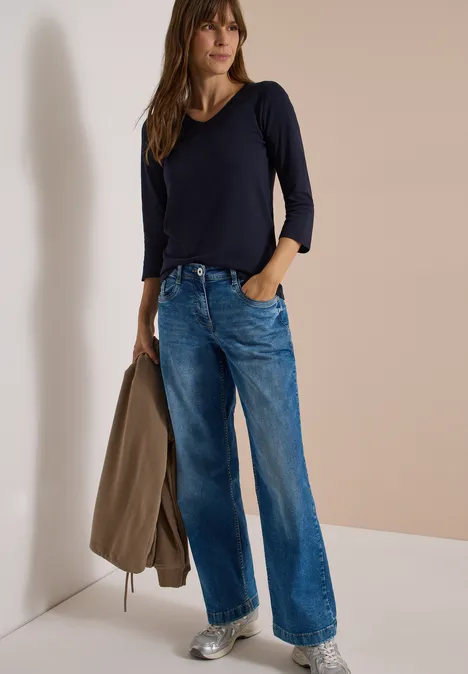 3/4-Arm Shirt in Unifarbe mit V-Neck urban dark blue Frau im dunkelblauen Longsleeve und weiten Jeans, hält eine Jacke locker in der Hand.