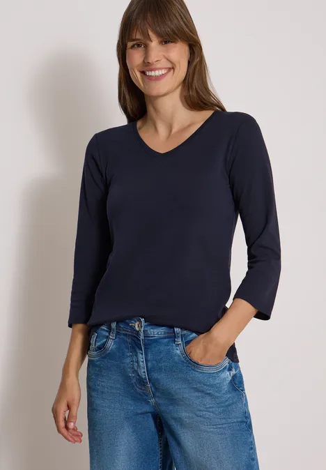 3/4-Arm Shirt in Unifarbe mit V-Neck urban dark blue Frau mit braunen Haaren trägt dunkelblaues Langarmshirt und blaue Jeans, lächelnd vor weißem Hintergrund.