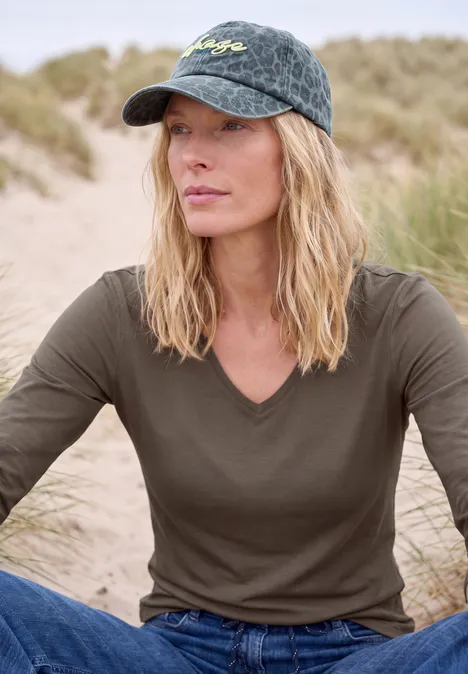 Femme en casquette et tee-shirt vert, assise détendue dans un paysage de dunes.
