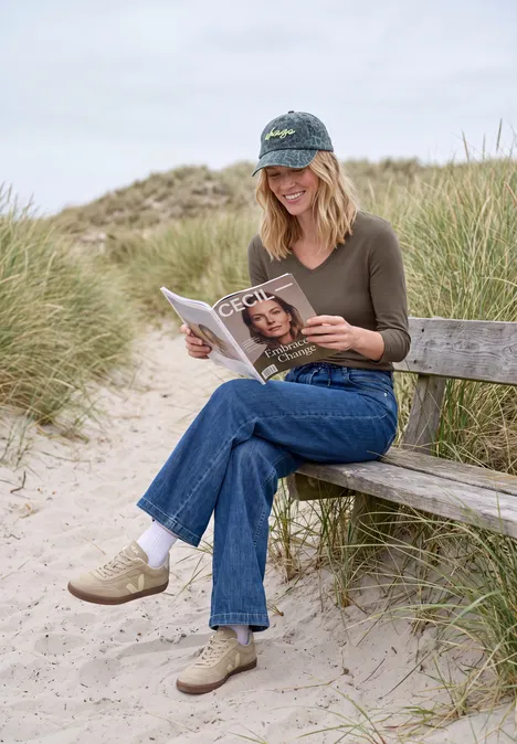 Une femme assise sur un banc entre les dunes au bord de la mer lit un magazine.
