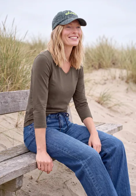 Femme souriante en jean et casquette assise sur un banc de plage, herbes de dune en arrière-plan.