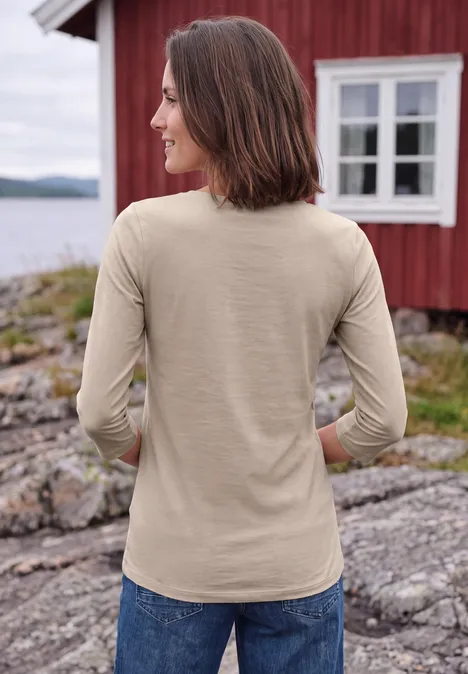 Femme en haut beige et jean debout sur des rochers devant une maison rouge au bord de l'eau