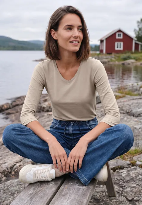 Une femme souriante assise en tailleur au bord du lac, avec une maison rouge en arrière-plan.