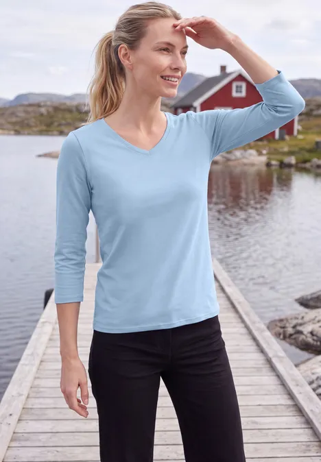 3/4-Arm Shirt in Unifarbe mit V-Neck cosmopolitan blue Frau in blauer Bluse steht auf Holzsteg vor See und Haus im Hintergrund