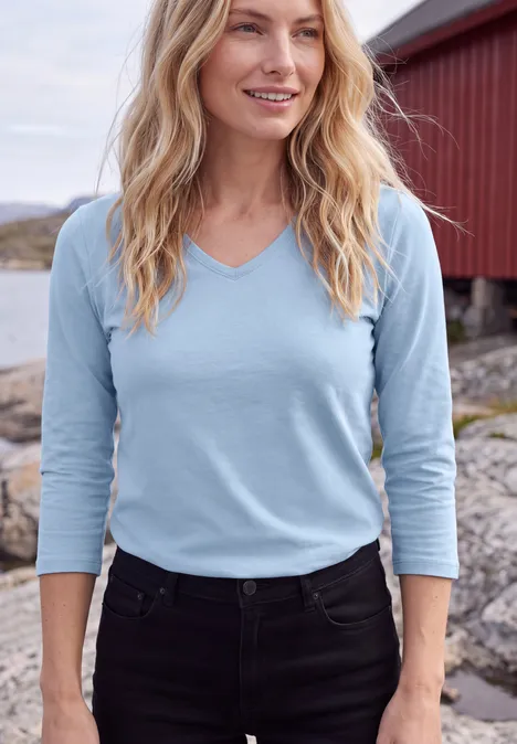 3/4-Arm Shirt in Unifarbe mit V-Neck cosmopolitan blue Eine blonde Frau mit hellblauem V-Ausschnittshirt und schwarzer Hose vor roter Holzhütte am Wasser.
