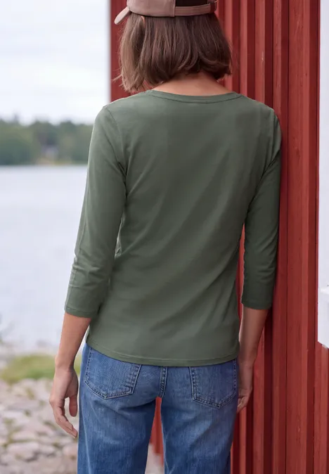 Frau von hinten in grünem 3/4-Langarmshirt und Jeans steht an roter Holzwand am See.