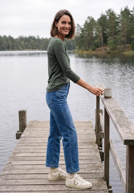 Eine lächelnde Frau in grünem Oberteil und blauen Jeans steht auf einem Steg am See und hält eine Tasse.
