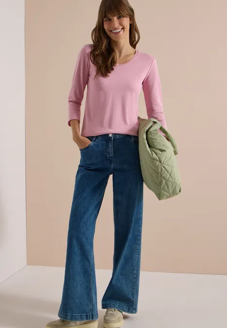 3/4-Arm Shirt in Unifarbe mit V-Neck mellow rose Frau in pinkem V-Pullover, weitem Jeans, weiße Sneaker, hält gesteppte grüne Jacke.