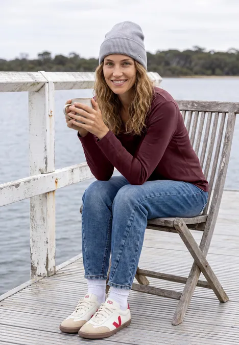 Une femme souriante est assise sur un quai, tenant une tasse, portant bonnet, pull et jean.