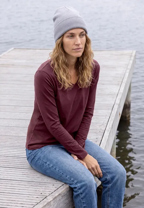 Femme en bonnet gris, haut bordeaux et jeans, assise pensivement sur une jetée en bois au bord de l'eau.