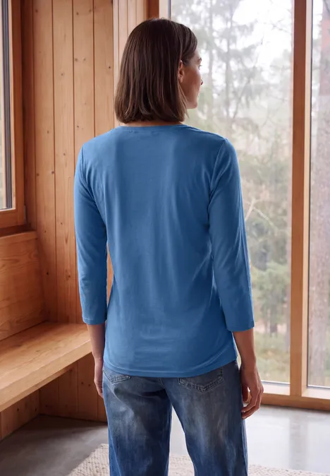 Frau von hinten in blauem Shirt und Jeans, steht in hellem Holzraum vor großen Fenstern mit Blick auf Wald.