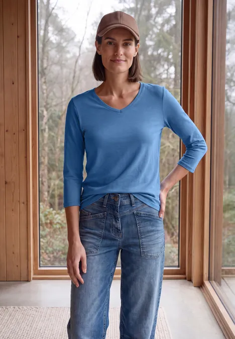Frau in blauer V-Neck-Strickbluse, Jeans und Cap steht lächelnd vor raumhohen Fenstern aus Holz.