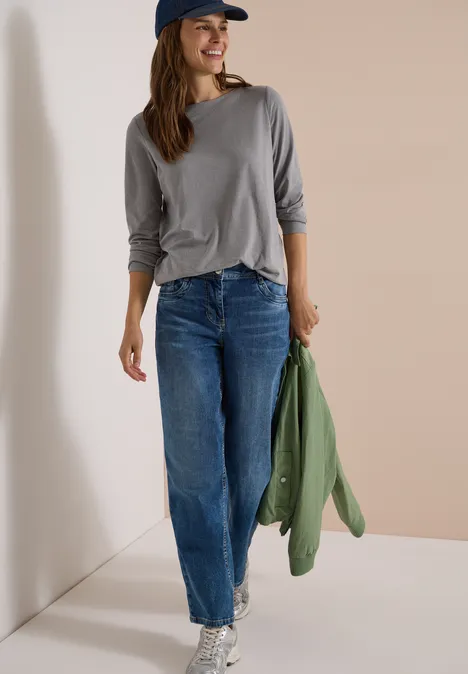 Eine Frau in grauem Shirt, blauen Jeans und Baseballcap lehnt entspannt an einer Wand und hält eine grüne Jacke.