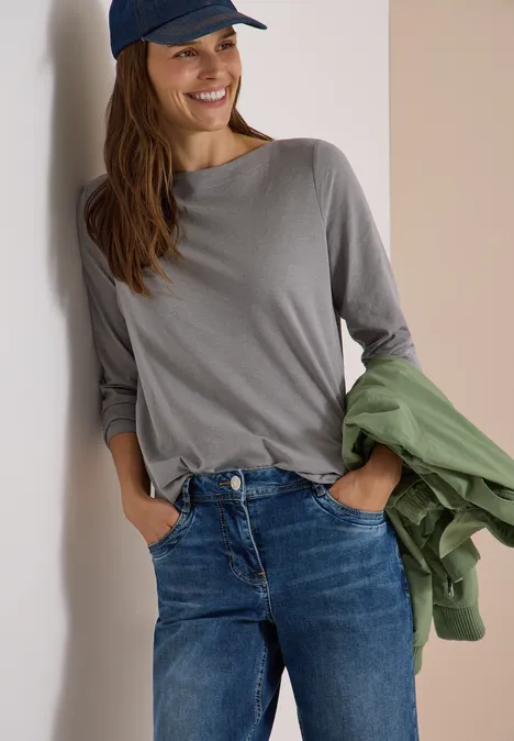 Frau lehnt lächelnd an Wand, trägt Jeans, graues Shirt, blaue Cap und hält grüne Jacke.
