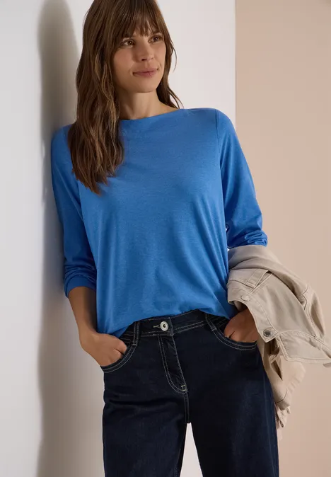 Frau lehnt lässig an Wand, trägt blauen Pullover, Jeans und hält eine Jacke in der Hand.
