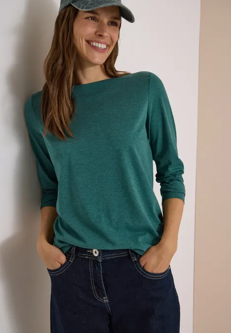 Langarm Shirt mit U-Boot-Ausschnitt dark fir green melange Frau in grünem Langarmshirt, Jeans und Cap lehnt lächelnd an Wand, Hände in den Hosentaschen.