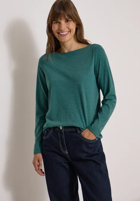 Langarm Shirt mit U-Boot-Ausschnitt dark fir green melange Eine lächelnde Frau in grünem Langarmshirt und blauen Jeans vor hellem Hintergrund.