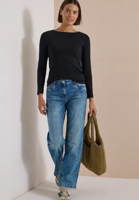 Langarm Shirt mit U-Boot-Ausschnitt Black Junge Frau in schwarzem Langarmshirt und blauen Jeans mit grüner Tasche lehnt lächelnd an heller Wand.