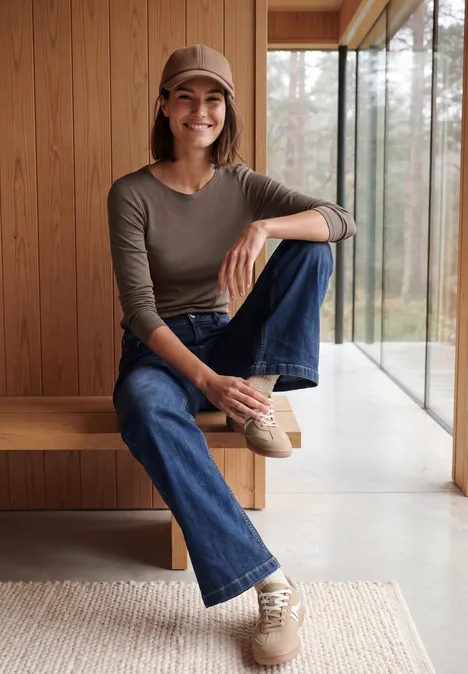 Frau in Jeans, Pullover und Cap sitzt lässig auf einer Holzbank vor großen Panoramafenstern.