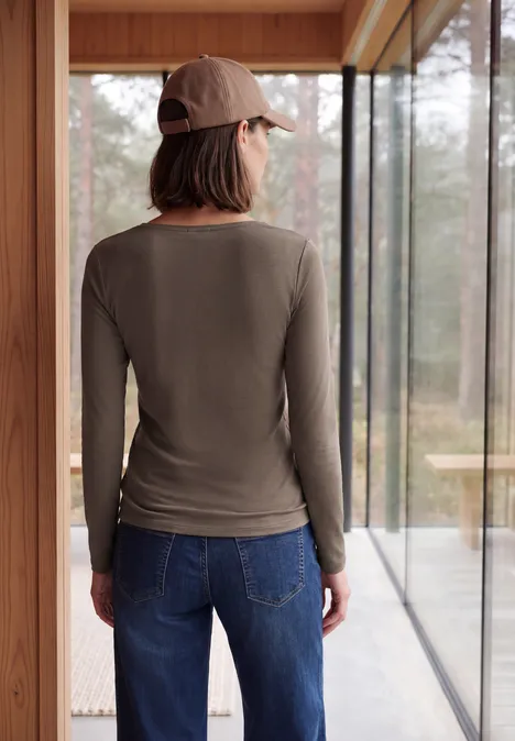Frau von hinten in lässigem Langarmshirt, Jeans und Baseballcap, stehend vor großem Fenster mit Waldblick.