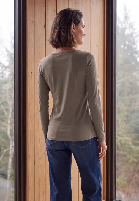 Eine Frau steht vor einem Fenster, Rückenansicht trägt ein olivgrünes Langarmshirt und blaue Jeans.