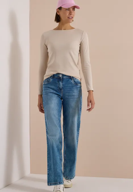 Langarm Shirt mit U-Boot-Ausschnitt cashmere beige Eine junge Frau mit rosa Cap, beigem Langarmshirt, weiten blauen Jeans und weißen Sneakern lehnt lässig an einer Wand.