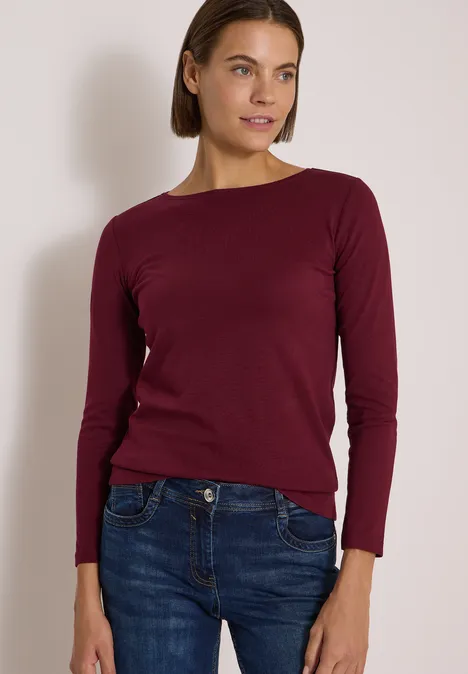 Langarm Shirt mit U-Boot-Ausschnitt red wine Eine Frau trägt einen dunkelroten Pullover und blaue Jeans, steht vor einer hellen Wand.