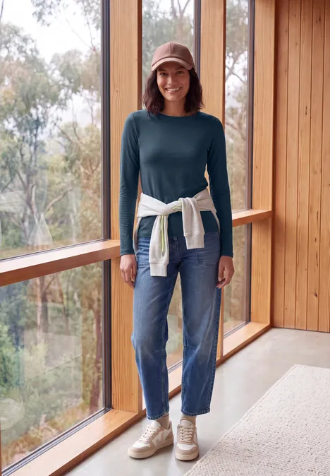 Langarm Shirt mit U-Boot-Ausschnitt moonlight blue Frau in Jeans, grünem Pullover, Cap und Sneakers steht in hellem Holzraum am Fenster.