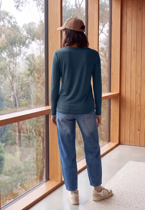 Langarm Shirt mit U-Boot-Ausschnitt moonlight blue Frau mit Cap, langärmeligem Shirt und Jeans schaut aus großem Fenster auf bewaldete Umgebung.