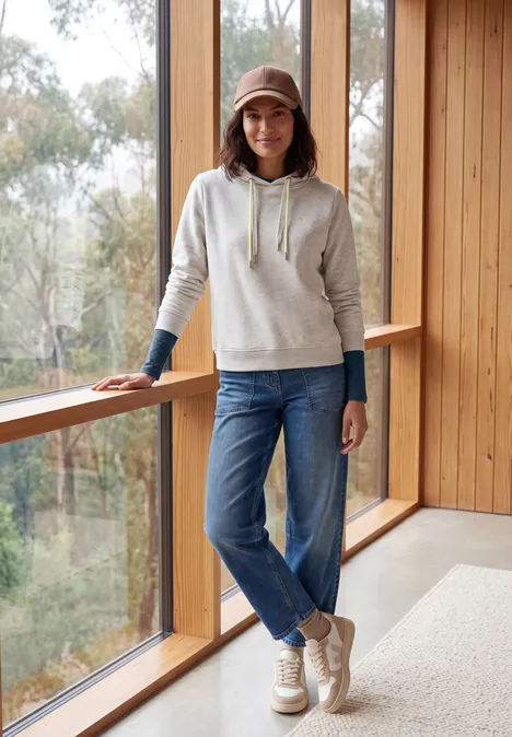 Langarm Shirt mit U-Boot-Ausschnitt moonlight blue Junge Frau mit Hoodie, Jeans und Cap lehnt an einem Holzgeländer vor großen Fenstern.