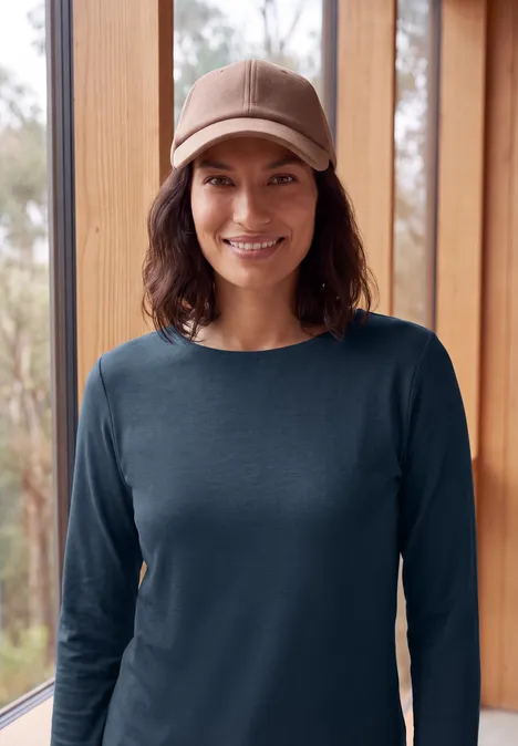 Langarm Shirt mit U-Boot-Ausschnitt moonlight blue Eine lächelnde Person in einem dunkelblauen Pullover und Schirmmütze vor großen Holzfenstern.