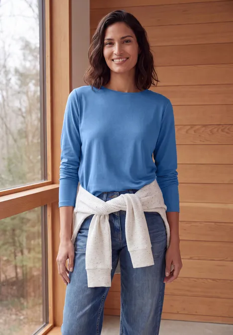 Langarm Shirt mit U-Boot-Ausschnitt north blue Eine lächelnde Frau in blauem Pullover und Jeans, mit weißem Sweatshirt um die Hüfte, in einem hölzernen Raum.