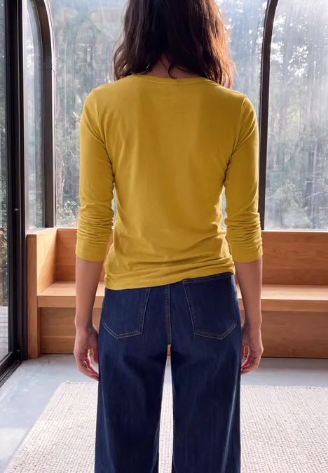 Frau steht in gelbem Oberteil und blauer Jeans vor einem großen Fenster.