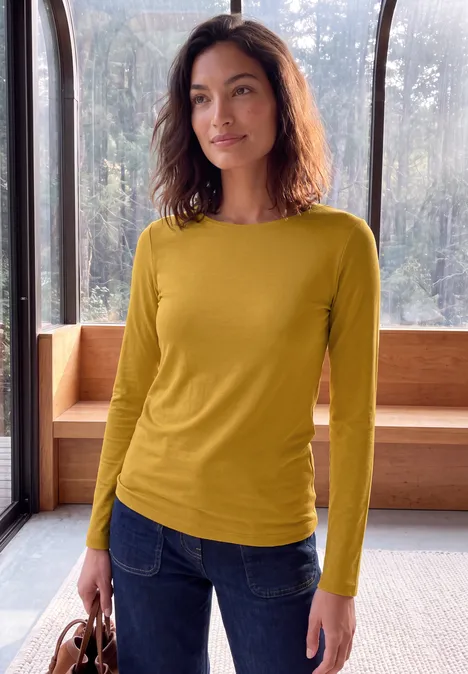 Eine Frau trägt ein gelbes Langarmshirt und blaue Jeans, steht mit Tasche in modernem, hellem Raum.